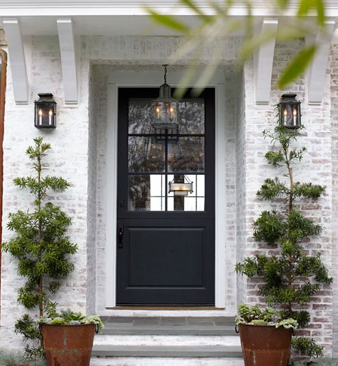 Entrace to a white washed brick house with rusty metal planters on either side, stone stairs, a black front door, and lantern style lights