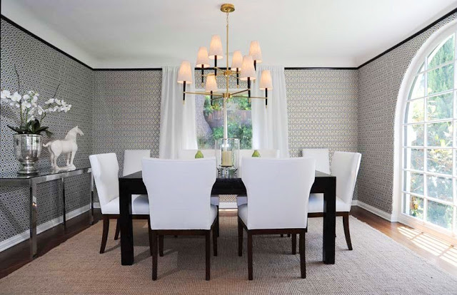 Dining room with graphic wallpaper, black crown molding, a black table surrounded by white chairs, a brass chandelier, and an arched picture window