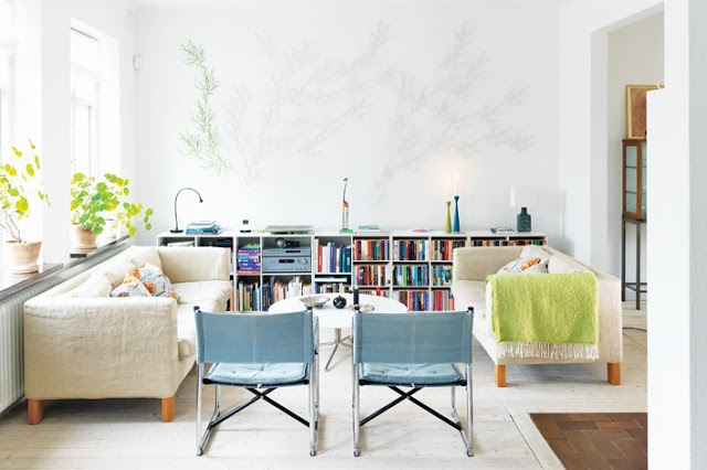 Living room with dueling white sofas, two chairs with blue cushions, a wood floor and a wall length bookcase stuffed full of books