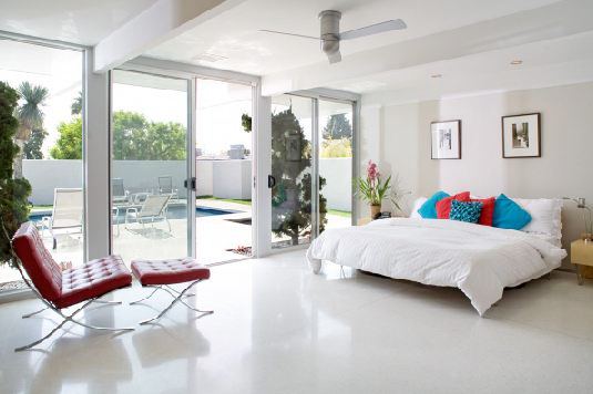 White bedroom with tile floor, red and blue accent pillows and a red barcelona chair with matching ottoman. There is a large sliding glass door that leads out to the pool