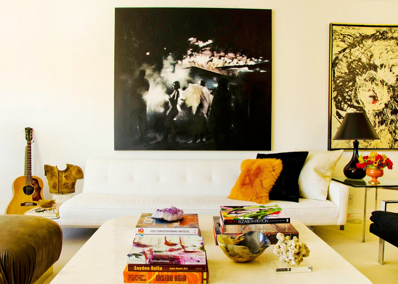 Living room with white sofa with gold, black and white accent pillows, a white coffee table, glass side tables, a leather ottoman and modern art on the walls