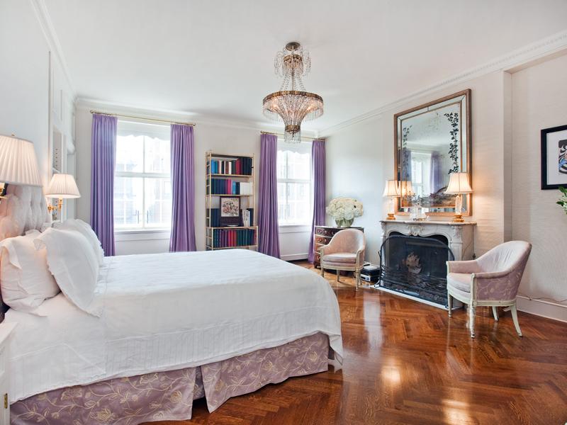 Master bedroom in a park avenue apartment with purple floor length curtains, a marble fireplace with a large vintage mirror on the mantel, a crystal chandelier and herringbone wood floor