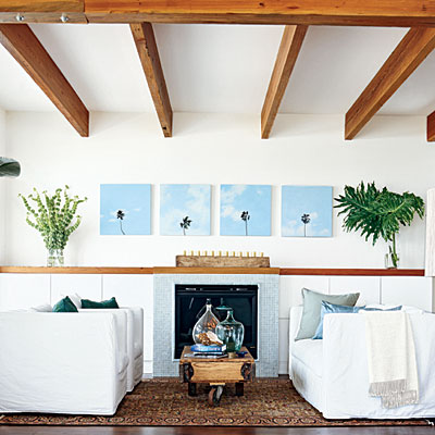Living room with white walls and exposed ceiling beams, wood floor, white armchairs and sofa with blue accent pillows, a fireplace with blue tile surround and on the wall above the fireplace are four paintings of the tops of palm tress bookended by a glass vase of palm leaves