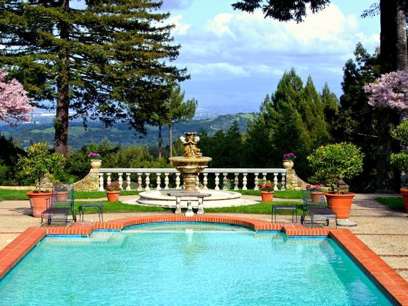 Pool in the backyard of a florentine villa overlooking a gorgeous mountain side with a gold fountain, bricks surrounding the pool and a small lawn