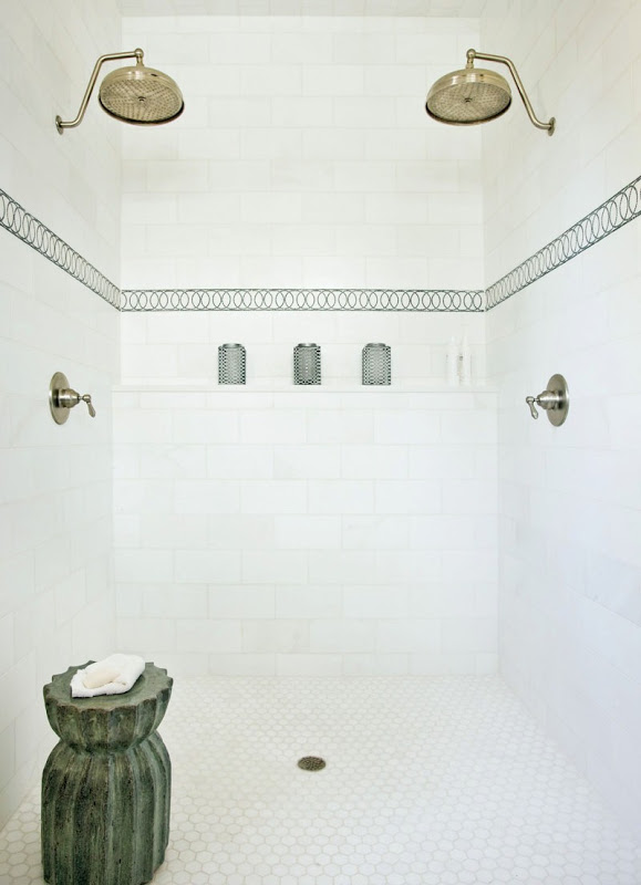 Shower white tile tile on the walls with a patterned accent stripe tile, two gold shower heads, hexagon tiles on the floor and a stone towel holder