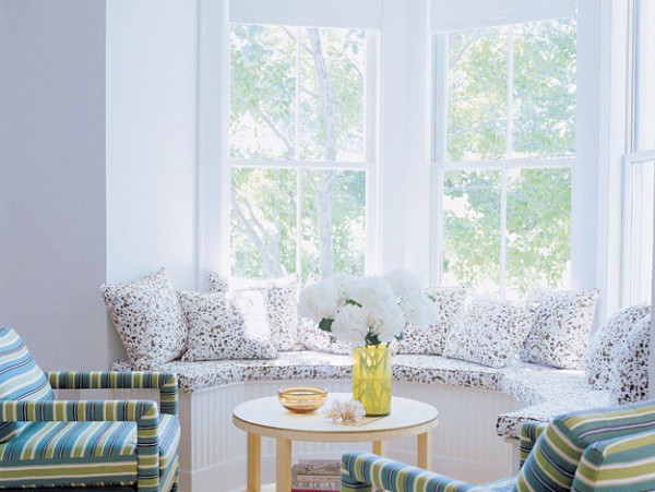 Sitting room with a white window seat with lacy cushions, a round wood table holding white hydrangeas in a yellow vase and two bright green, blue and turquoise striped armchairs