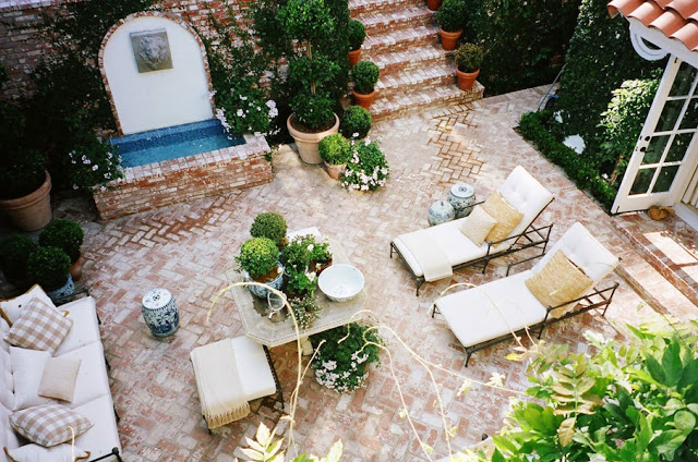 ariel view of a brick lined outdoor patio with metal lawn chairs with neutral cushions, a matching sofa, a small brick fountain and lots of potted plants