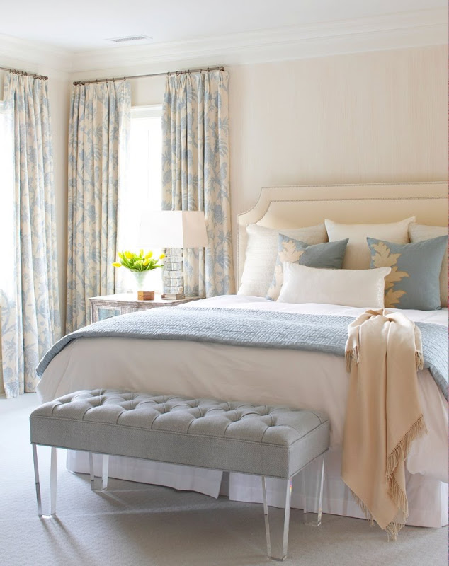 Bedroom in a cottage with blue and cream floor length curtains, a blue tufted ottoman at the foot of the bed, a cream upholstered headboard with blue, white and cream accent pillows 