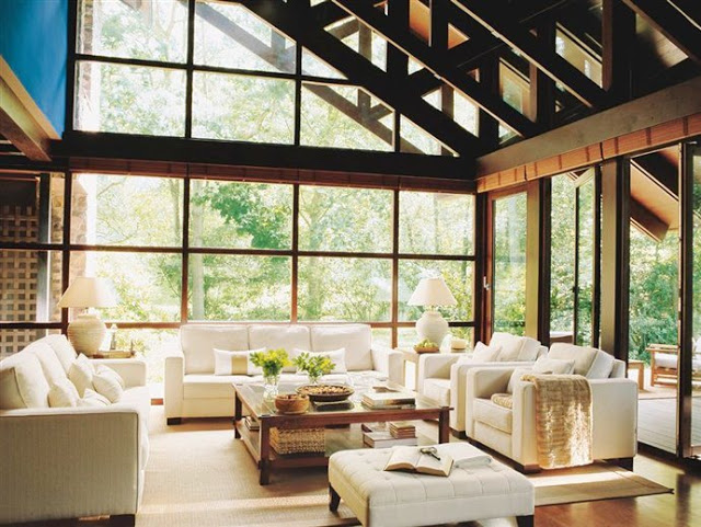 Living room in a converted mill with the walls replaced with glass windows and doors, white sofas and arm chairs and a wood coffee table 