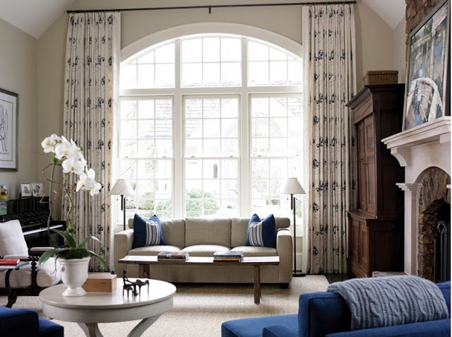 Living room with high ceiling, large arched window with floor length printed drapes, a light grey sofa, blue armchairs, a piano, a wooden chest of drawers and a fireplace with an arched mantel