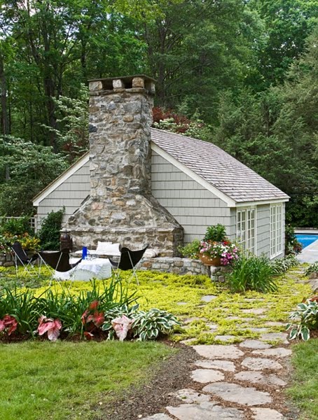 Exterior of a pool house with a stone chimney, butterfly chairs arranged around a small table and the stone path through the grass leading up to this scene