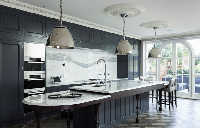 Kitchen in a suburban London home with dark grey cabinets, marble counter tops and backsplash, stainless appliances, silver pendant lights, carved crown moulding, ceiling medallions, parquet wood floors and leather barstools