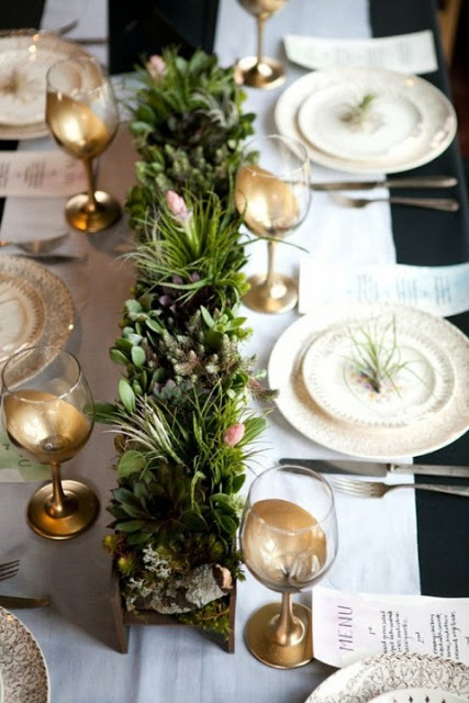 Holiday table setting with gold wine glasses and green plants in a gold rectangular vase separating the two sides of the table