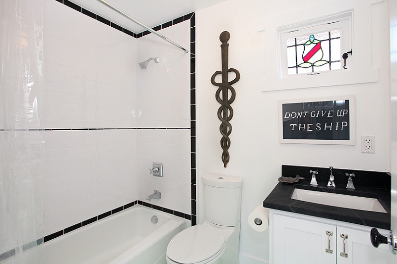 Bathroom in a cottage with white subway tile with a black border in the shower, white cabinets and a black countertop. On the wall above the sink is a framed piece that says "Don't Give Up The Ship"