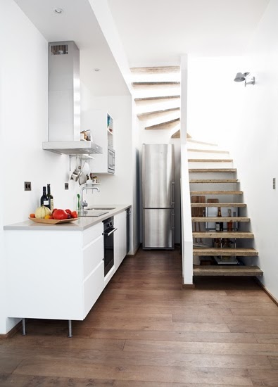 Small white kitchen under a staircase with a tall narrow fridge and stainless hood