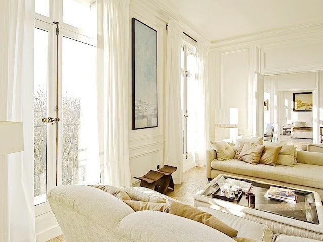 living room with high ceilings, floor length white curtains, neutral dueling sofas, molded walls and a glass coffeetable