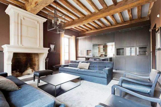 den with white molded fireplace, dark wood floors, exposed beams, dark blue dueling sofas separated by coffee table, a light gray rug, and dark wood cabinets 