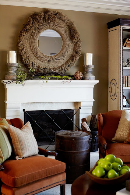 close up of fireplace with woven circle mirror and two candlesticks on mantel, diamond printed gate, a barrel converted into a side table and two orange chairs with neutral accent pillows