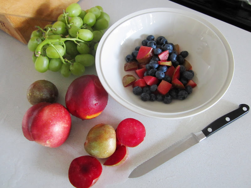 Pluots, nectarines, white nectarines and grapes are on a white kitchen counter with a knife. There is a white bowl on the same table with blueberries and some cut nectarines