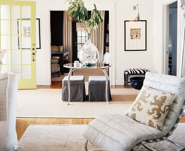 white entry way with a light yellow door, wood floor, a sea grass rug, a round mirror table holding a large tear drop vase with a plant, two grey ottomans are under the table, in the foreground there is modern white chair with a curved back