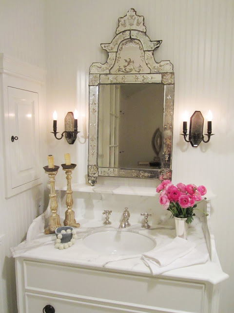 Bathroom in the House of Windsor with white beadboard paneled walls, marble counter top, white cabinets, a vintage etched mirror, two candle sticks, pink peonies and two wall mounted candle stick holder inspired lights