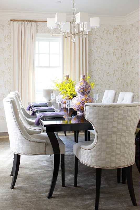 Dining room with floor to ceiling wallpaper, black polished table, floor length cream curtains and white armchairs with nail head trim