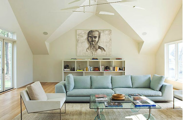 Living room with long light blue sofa, a white arm chair, glass coffee table, wood floor, a white cubby style book shelf with a large portrait above it