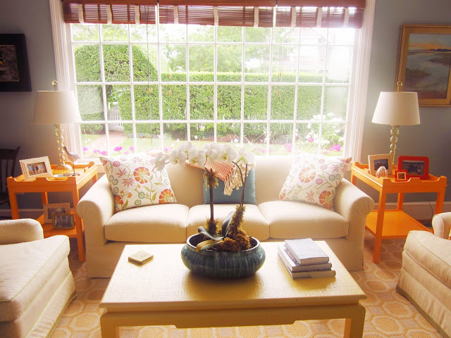 paned windows with bamboo shades, two yellow accent tables match the coffee table, a neutral sofa with blue and flower print accent prints. 