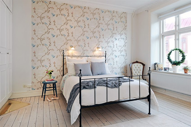 Bedroom in a Swedish apartment with iron bed frame, light knotty wood floor, an upholstered armchair, wall mounted lights, wall covered in a delicate wallpaper with a floral pattern with birdcages and a large window
