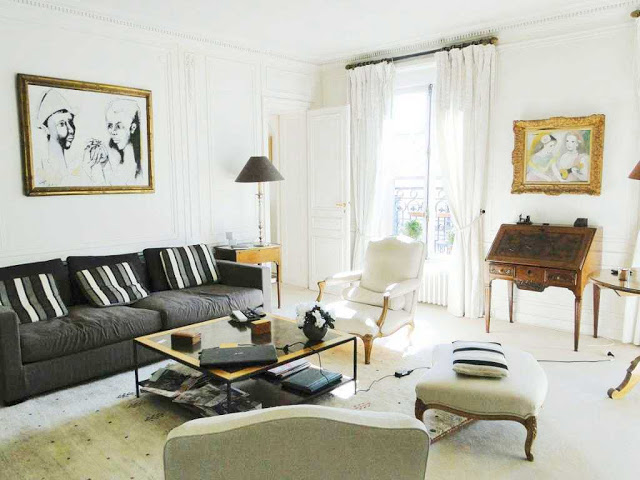 Den in a Paris apartment with a writing desk, dark grey sofa, light gray armchairs and ottoman, french doors and a square coffee table with metal legs