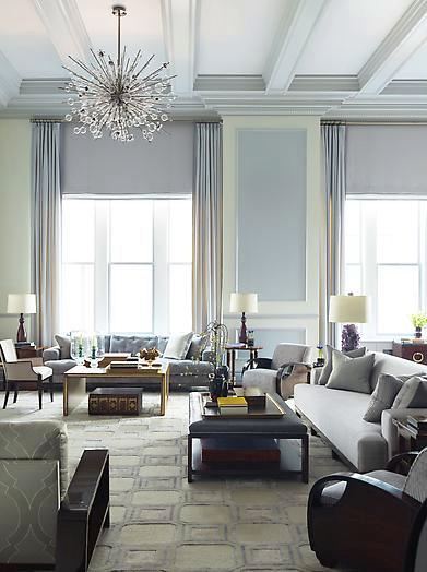 Living room by Steven Gambrel with high coffered ceilings, a modern spikey chandelier, grey-lavender painted walls to created a panel effect, floor length curtains, grey sofas and matching armchairs, a leather ottoman has been turned into a coffee table and a greyish lavender area rug covered in squares.