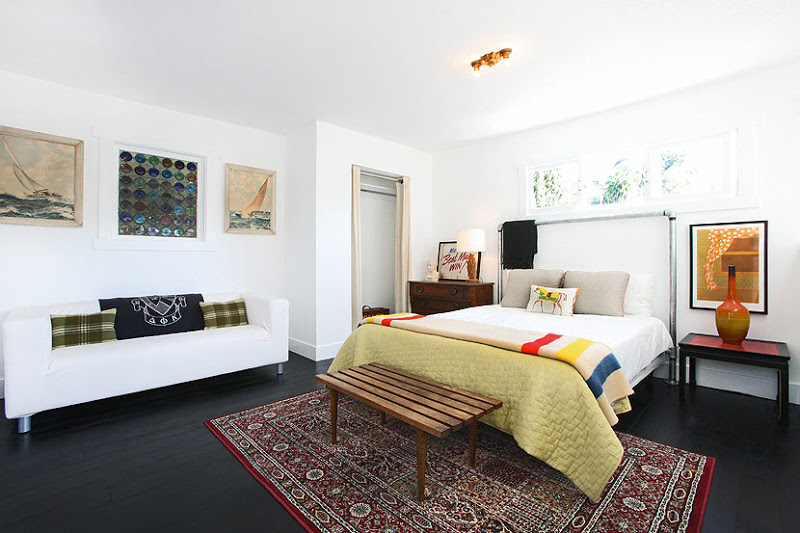 Bedroom in a cottage with stained wood floor, a red Moroccan rug, a white sofa with green plaid accent pillows and a navy throw with a seal on it, a bed with a metal headboard, a bench is at the foot of the bed and white walls