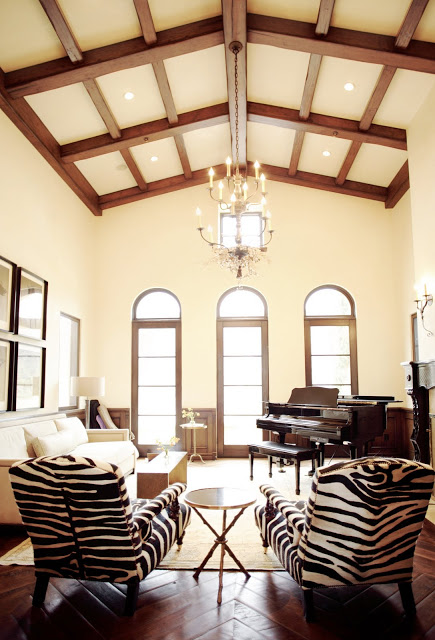 Living room with super high ceilings, exposed beams, arched windows, herringbone wood parquet floors, a piano, and very neutral furnishings except for two upholstered zebra print chairs