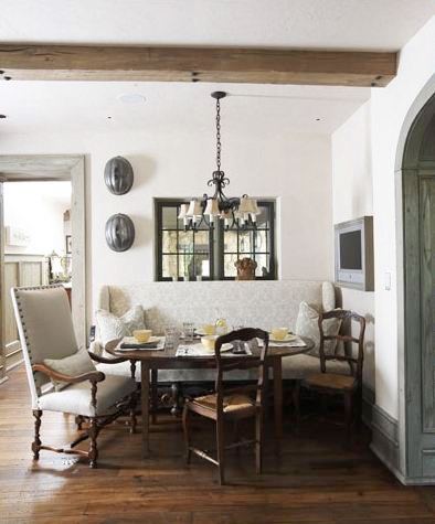 Breakfast nook with exposed beams, stained wood floor, a wire chandelier, bench style seating on one side of an oval table with two wood chairs with rope seats and an upholstered arm chair with nail head trim on the other