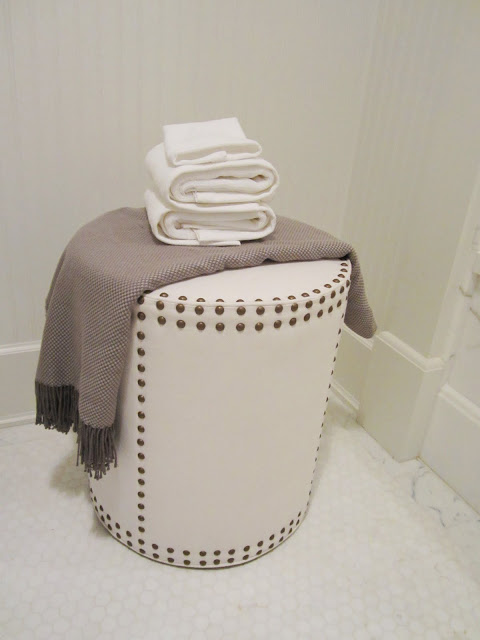 Close up of the little leather stool with nail head trim holding a fringed throw and white towels in a small bathroom and white hexagon tile floor in the House of Windsor
