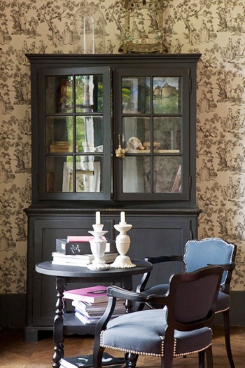A sitting area with classic Toile de Jouy wallpaper, two blue chairs white white nailhead trim around a small round table and a large cabinet with glass doors
