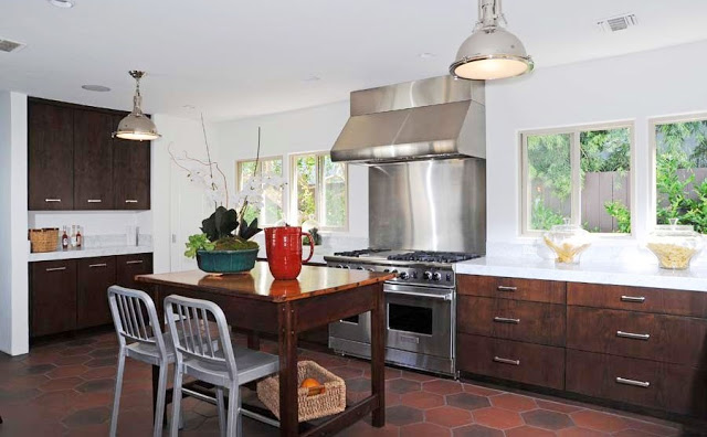 Kitchen with hexagon Saltillo tiles on the floor, stainless appliances, dark wood cabinets with silver drawer pulls and white counter tops and two silver pendant lights. Instead of an island, there is a wood table with two metal chairs
