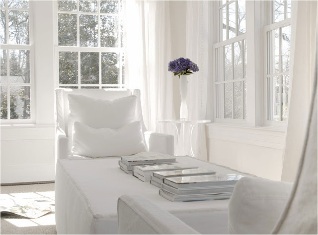 White room with a reading area surrounded by large encasement windows, dueling sofa chairs and books