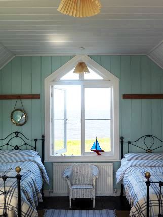 old fashioned iron twin beds with plaid bedding in a preppy beachy guest room with wood floor, a striped rug and a white wicker chair and a large pentagon shaped window overlooking an ocean or lake