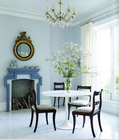 Soft blue dining room with a crystal chandelier, periwinkle carved wood geometric relief style mantel,round white Saarinen table with simple traditional wood side chairs, crown molding and a white painted wood floors