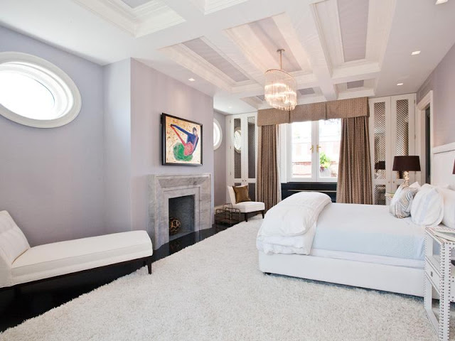 Master bedroom with coffered ceiling, black floor, large white shag rug, white chaise longue, lavender walls, a fireplace, chandelier, built in white cabinets with glass fronts, light brown curtains and oval windows