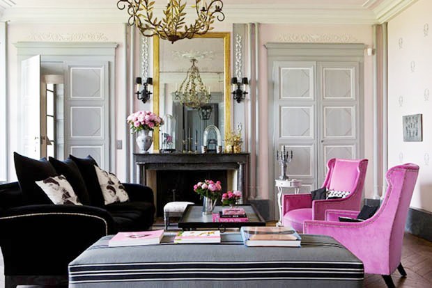 Pink and grey living room in a french mansion with black sofa, two pink wingback chairs, a marble fireplace wth a traditiona mirror, paneled doors, chandelier covered in leaves and a long grey ottoman