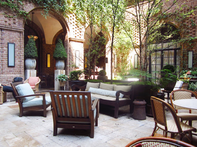 guest courtyard at the The Greenwich Hotel with stone floors, visible brick walls with arched entryways, potted plants and wooden and wicker arm chairs, tables and benches