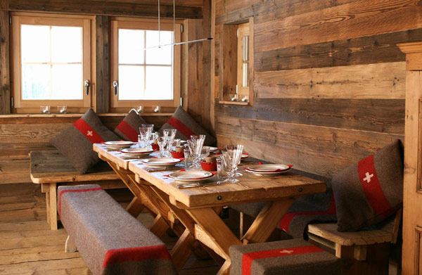 A dining room in a European ski chalet with rustic wood boards on the wall, rustic wood floor, bench style seats around a wood table with red and brown blankets and pillows