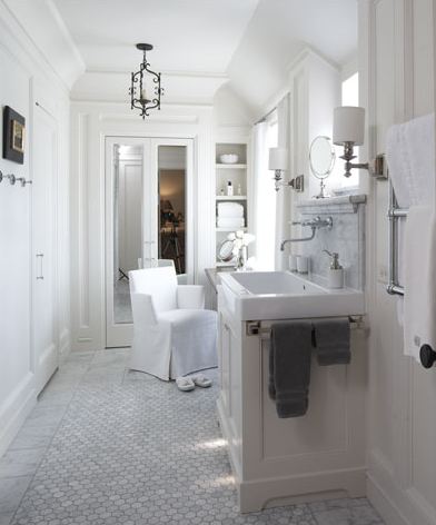 White bathroom with farmhouse style sink, a wall mounted faucet, marble hexagon Carrara marble tiles on the floor, a white armchair and a pair of white slippers