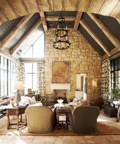 Rustic great rom in a tudor revival home with a stone fireplace, beamed ceilings, tile floor, a large area rug, large encasement windows with floor length patterned curtains, a wire chandelier, twin armchairs and an octagon side table
