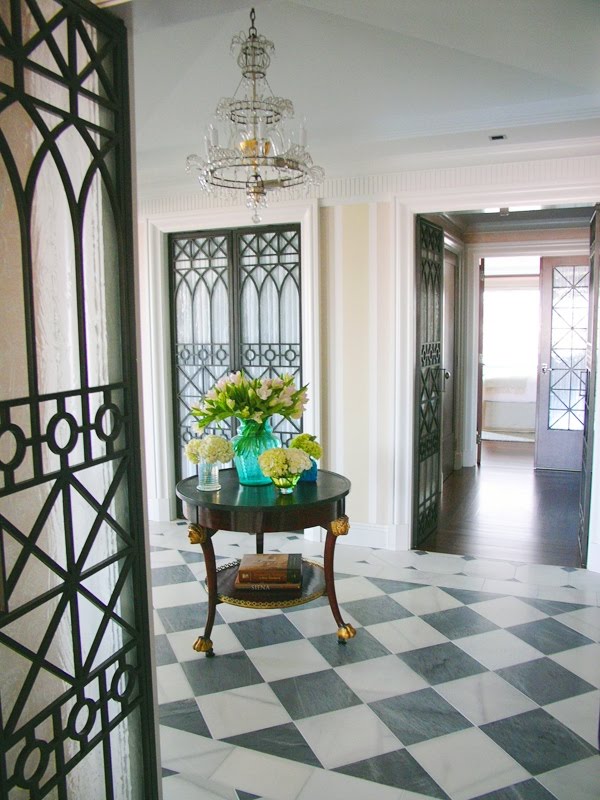 Foyer with checkered grey and off white marble floor, iron galss doors, crystal chandelier and a round table