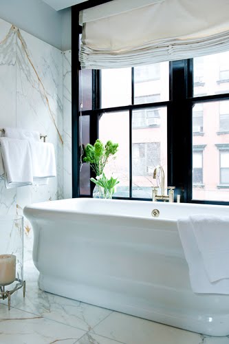 Bathroom with large white veiny marble slabs (probably a Calacatta gold marble), a stand alone porcelain tub, and glossy black trimmed windows 