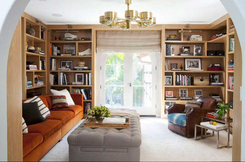 Library with orange wall length sofa, built in bookshelves full of books and other trinkets, a grey tufted ottoman, a brown leather armchair, white double doors with roman curtains on the far side of the room lead to a small patio