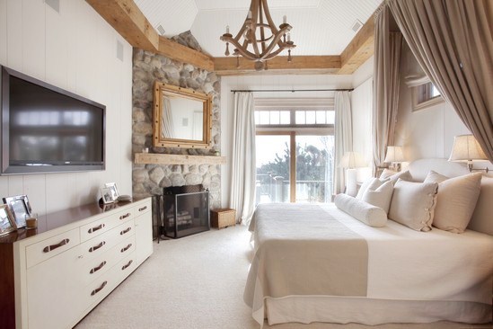 rustic master bedroom with exposed beams, stone fireplace with a large mirror in a wood frame on the mantel, canopy bed, wood chandelier and white drawers with wood handles